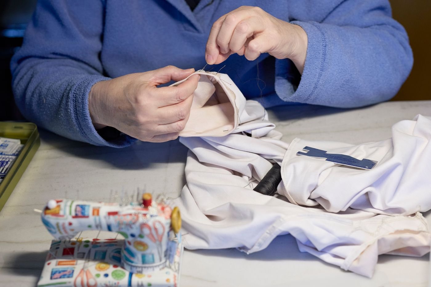 Representative close-up of a tailor's hands hand-sewing a white garment with needle and thread