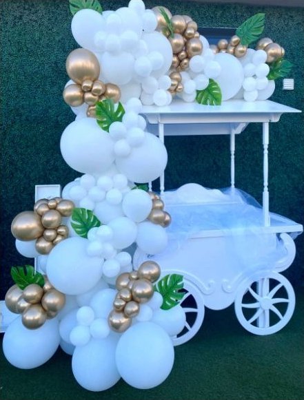 Representative photo of a white and gold balloon garland cascading down a candy cart against a greenery wall
