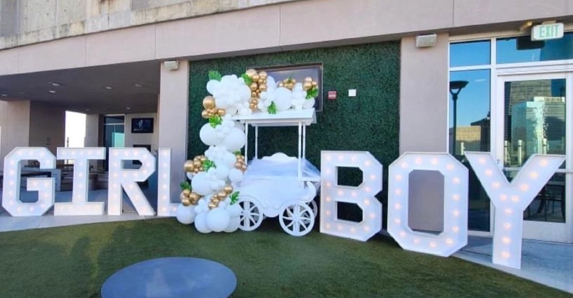 Representative photo of a gender-reveal setup by Bella'Loons: GIRL and BOY marquee letters with a white and gold balloon garland on a candy cart