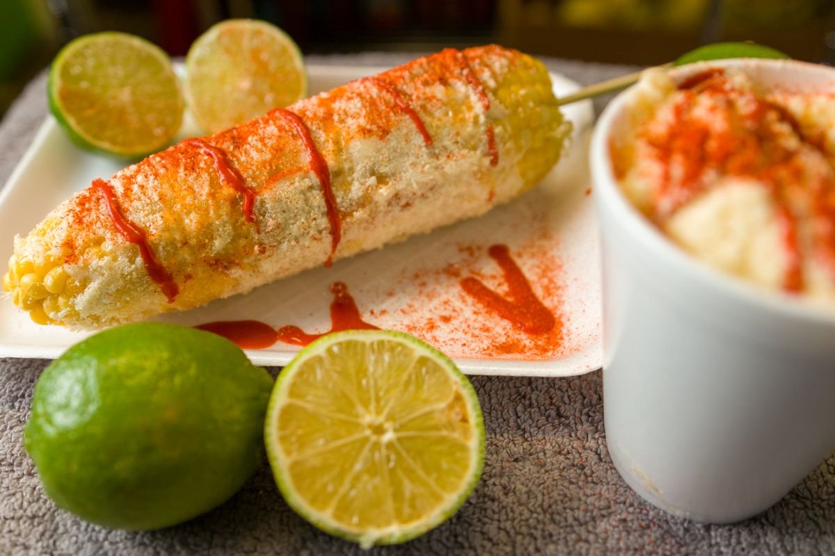 Representative photo of an elote entero on the cob with chile, lime and cotija, next to an esquite cup