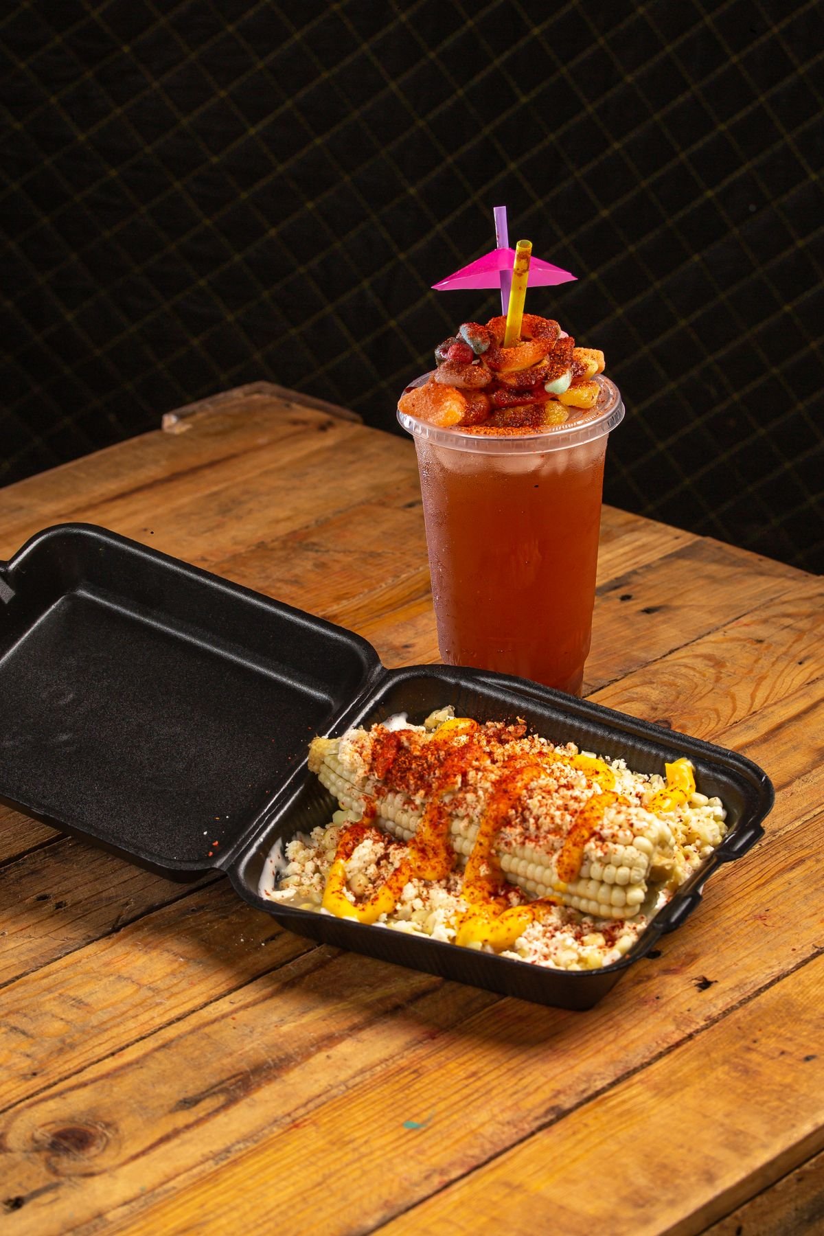 Representative photo of tostilocos with fruit and chile in a takeout box next to a fruit agua fresca