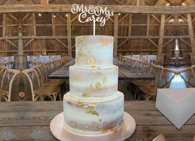 Three-tier semi-naked wedding cake with gold-leaf accents and a Mr & Mrs Carey topper, set up in a barn venue